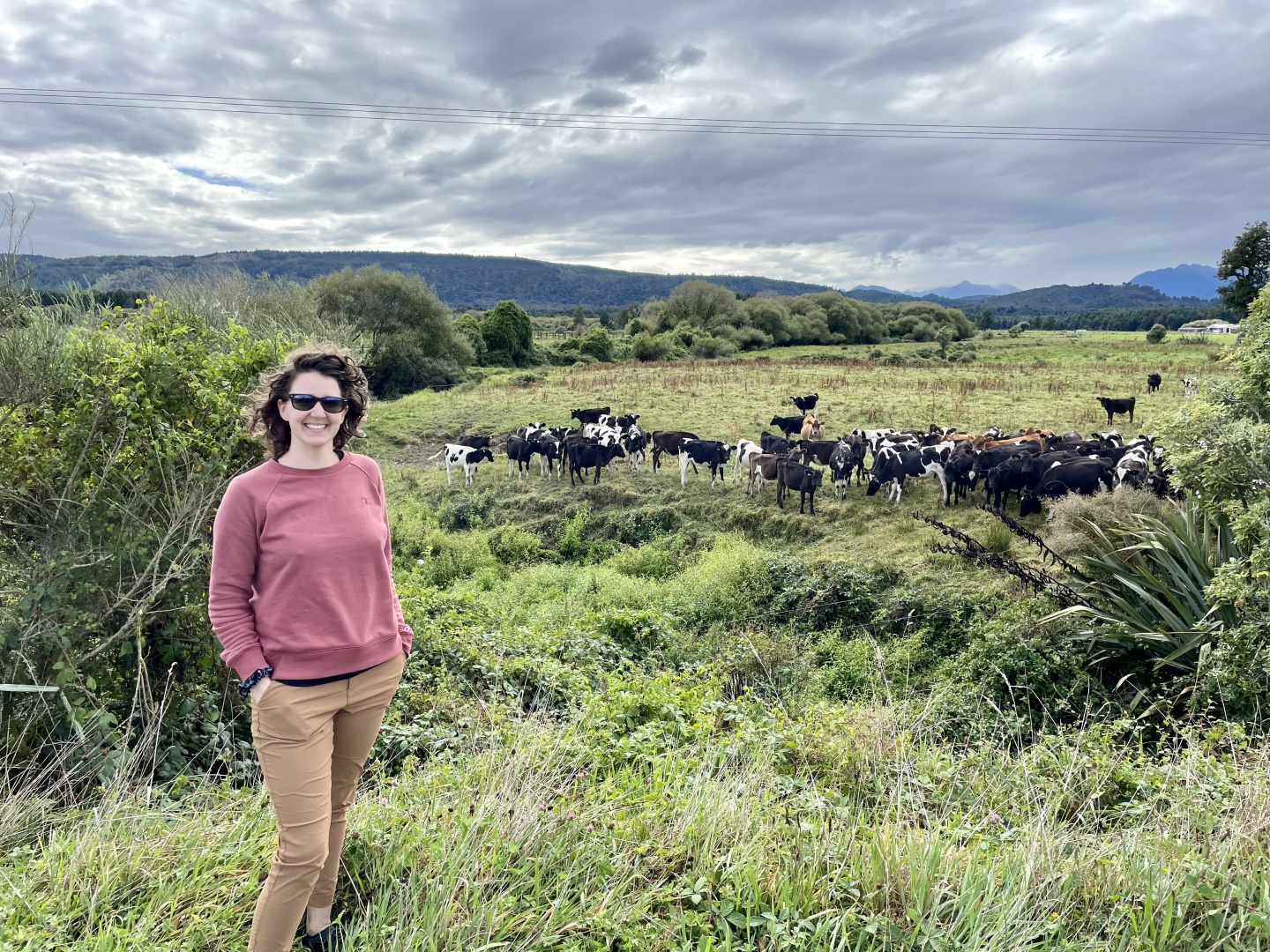 Kaitlyn smiling at the camera while standing in a field with several cows in the distance behind her.